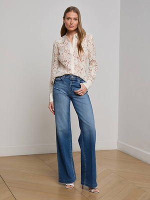 A woman with long straight hair stands indoors against a plain wall, wearing the L'AGENCE Maia Lace Blouse and high-waisted wide-leg blue jeans with white heeled sandals, one hand in her pocket.