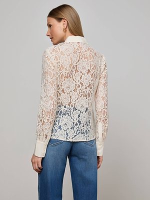 A woman models the Maia Lace Blouse by L'AGENCE in vintage white, featuring sheer floral lace, long sleeves, and collar with cuffs. She pairs it with blue jeans and faces away, showcasing the blouse’s delicate unlined pattern.
