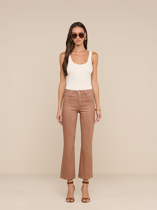 A woman with long brown hair, wearing sunglasses, a white sleeveless top, the Petite Kendra Coated Jean in light brown high rise style, and brown high-heeled sandals stands against a beige background.