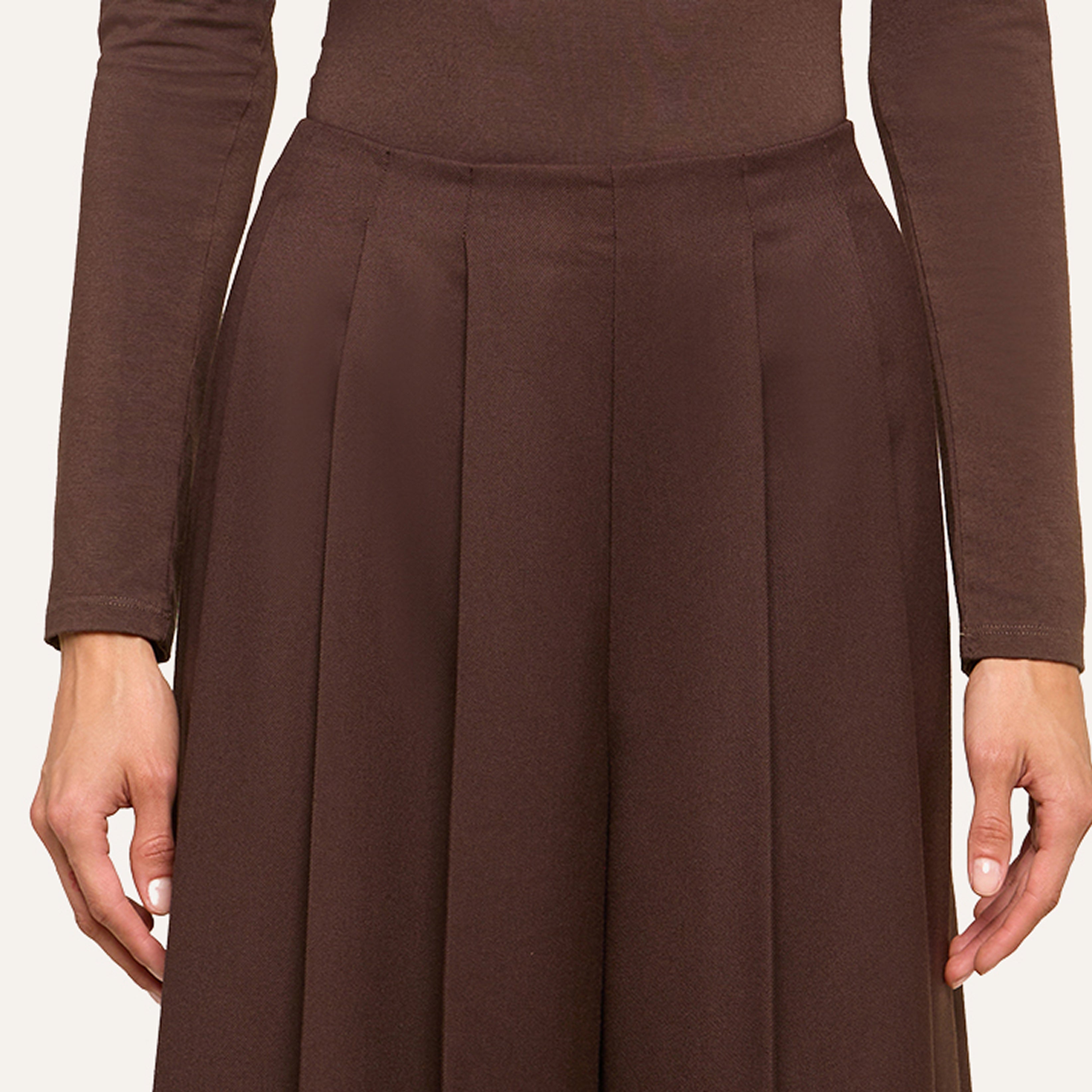 A person dressed in a long-sleeved brown top and a high-waisted pleated brown skirt, shown from shoulders to mid-thigh, exudes effortless style with both arms relaxed at their sides.