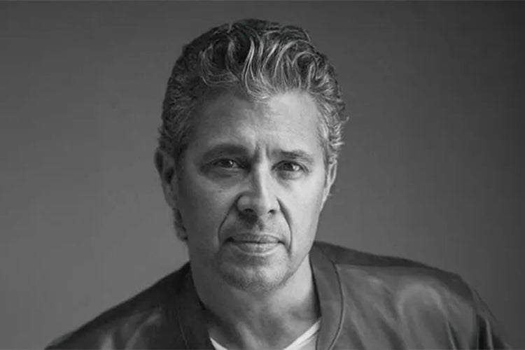 A man with short, wavy gray hair and light stubble looks at the camera with a neutral expression. He is wearing a dark jacket over a light shirt. The image is in black and white with a plain background.