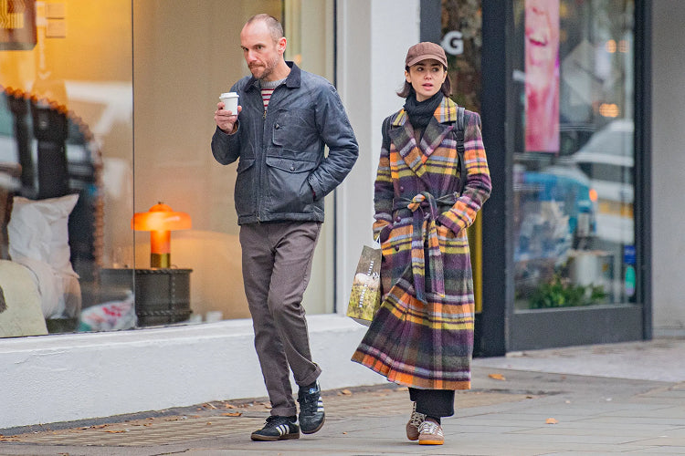 A man and a woman walk on a city sidewalk. The man, holding a coffee cup, wears a dark jacket and brown pants. The woman, in a colorful plaid coat and brown cap, carries a bag and looks ahead. Shop windows are visible behind them.