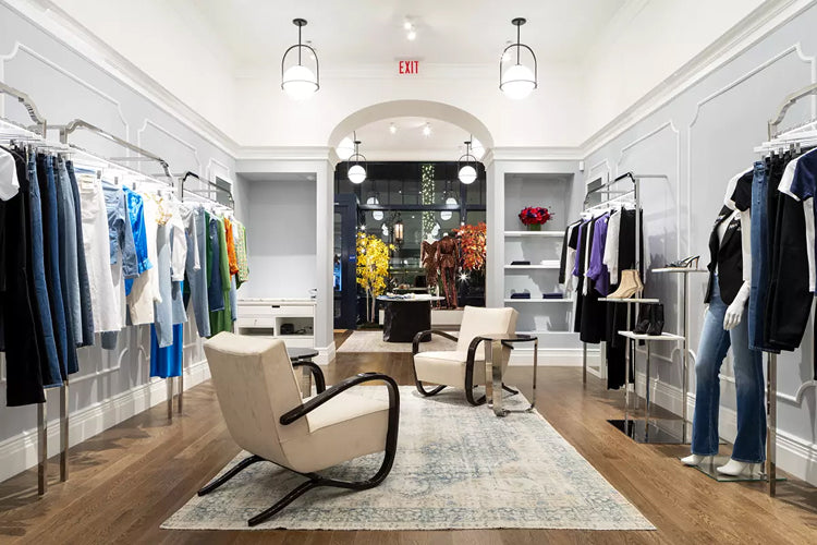 A modern clothing boutique with racks of colorful clothes on both sides, two cream armchairs around a rug in the center, mannequins on the right, and large windows at the back displaying plants and trees.