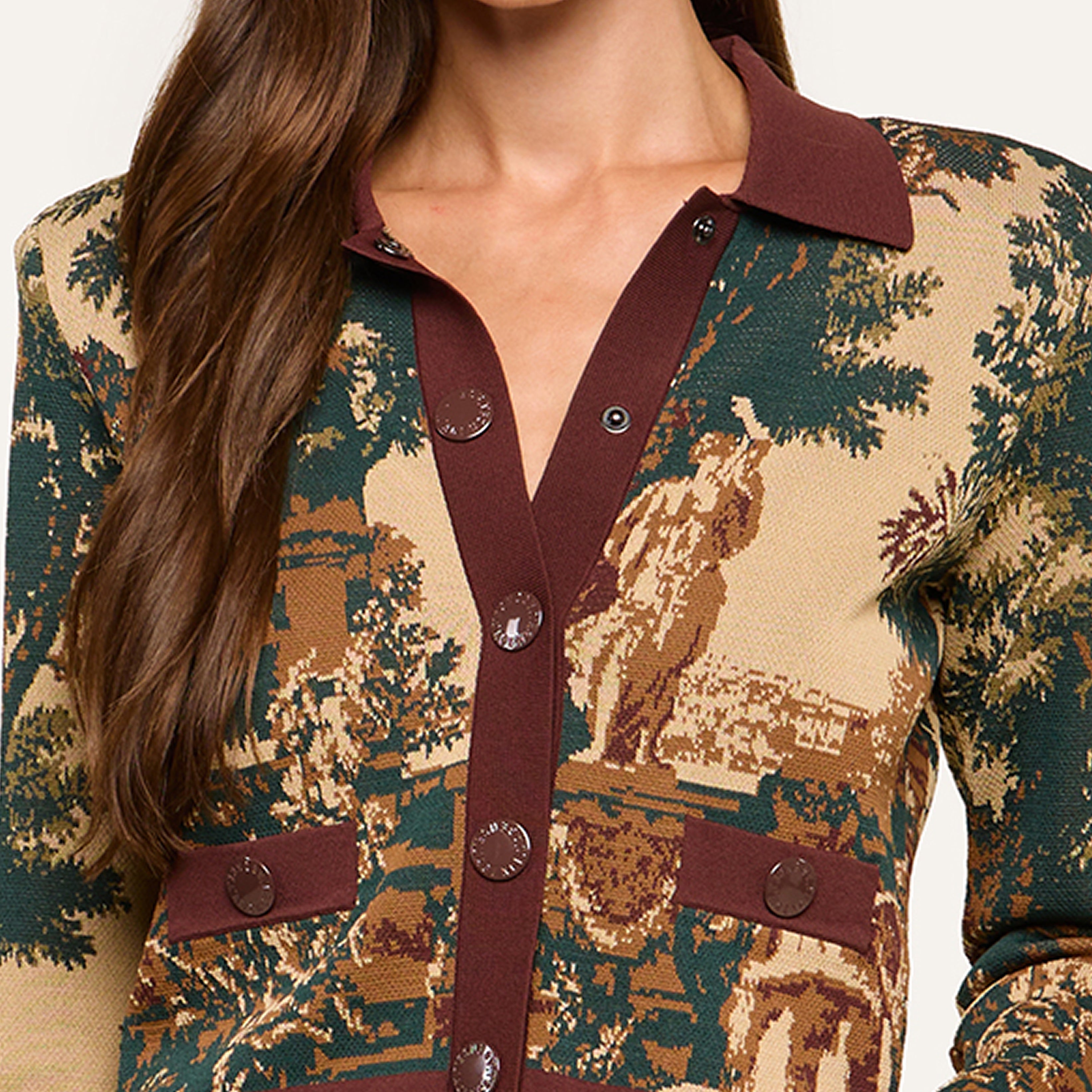 A woman wears a patterned jacket with brown buttons and collar, featuring a scenic tapestry design and two front pockets; her face is not visible.