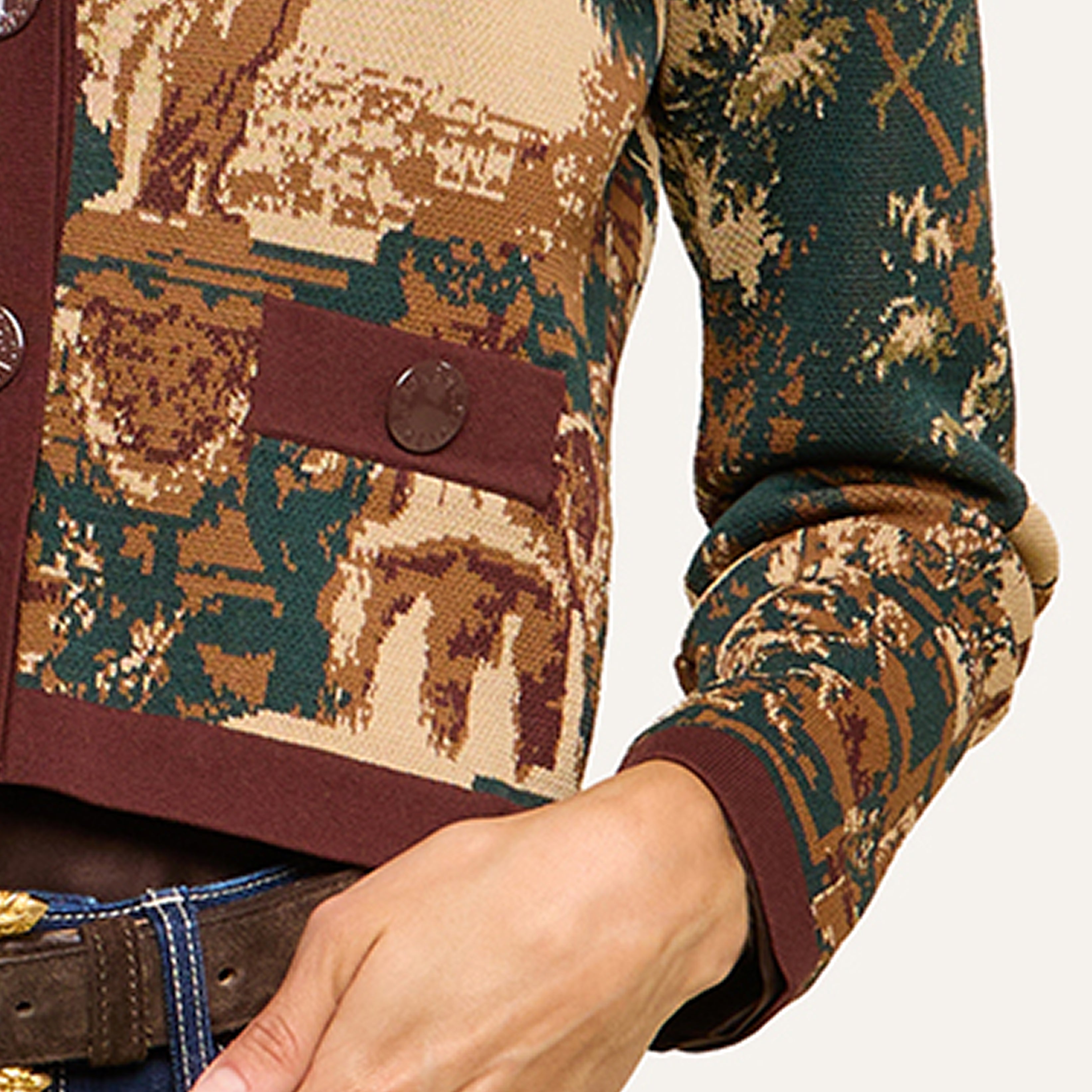 A close-up of a person wearing a knitted jacket with a scenic landscape pattern in brown, green, and beige, featuring two large buttons and rolled-up sleeves. The detailed knit showcases part of a hand and a belt.
