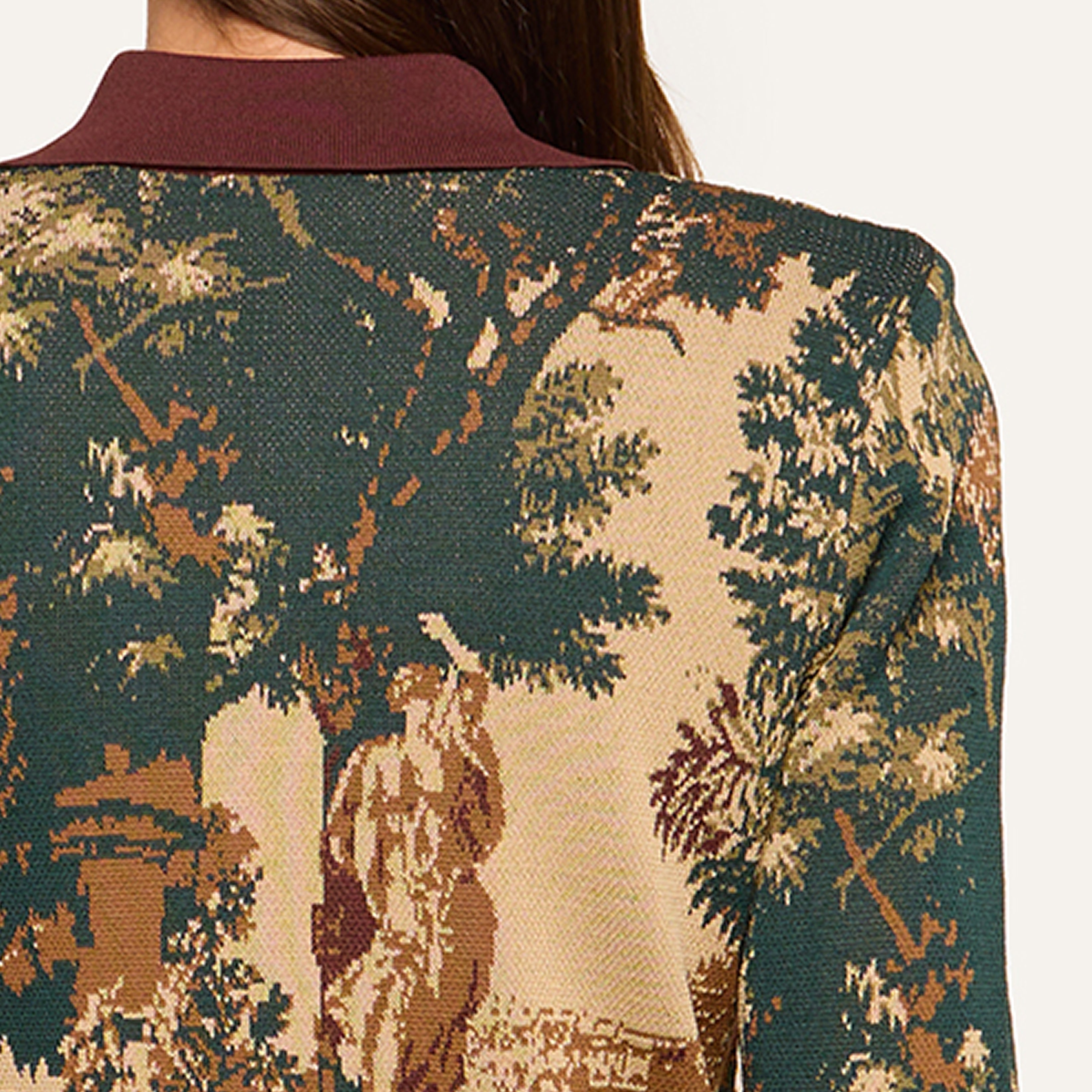 A close-up of the back of a jacket featuring a vintage tapestry design with trees and a classical scene, worn by someone with long brown hair.