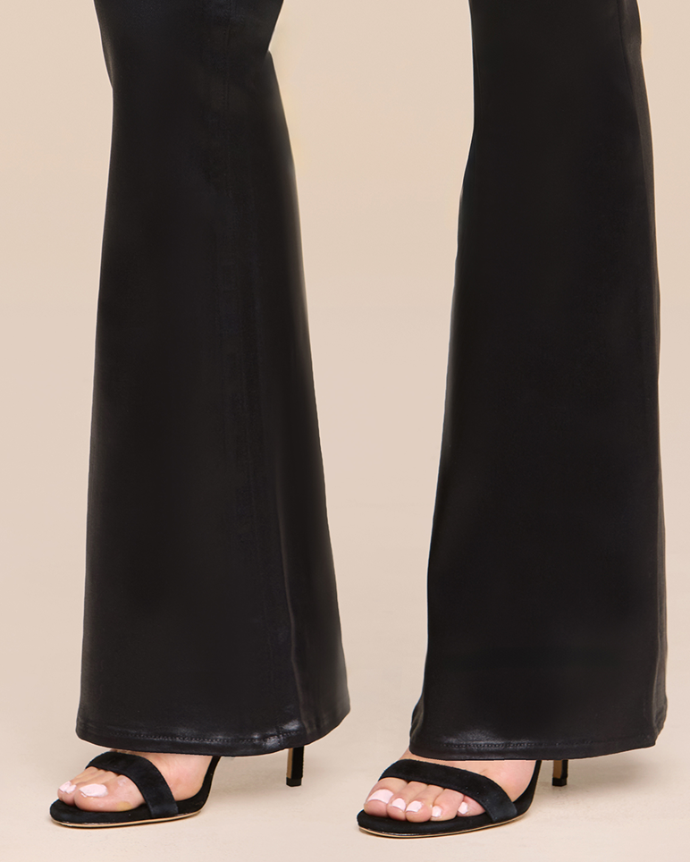 A person wearing black flared pants and black open-toe heeled sandals is standing on a beige background. Only the lower legs and feet are visible.