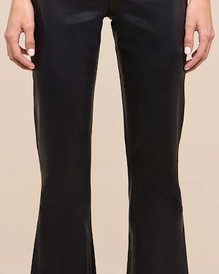 A person wearing black, shiny, flared pants stands with their hands relaxed at their sides against a beige background. Only the lower torso, legs, and hands are visible.