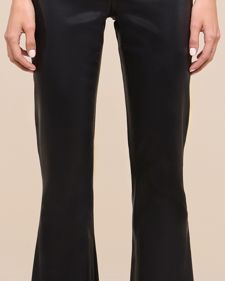 A person wearing black, shiny, flared pants stands with their hands relaxed at their sides against a beige background. Only the lower torso, legs, and hands are visible.
