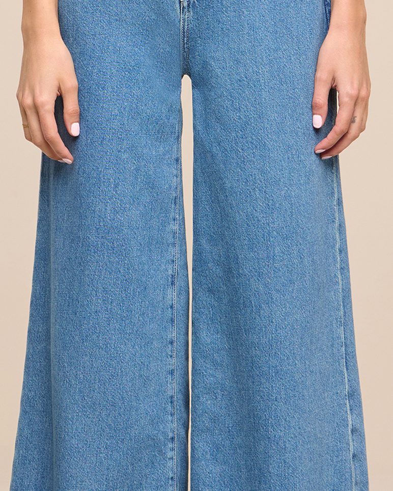 A person wearing high-waisted, wide-leg blue jeans stands with their hands relaxed at their sides against a beige background.