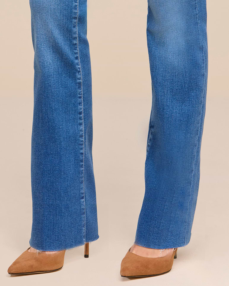 Person wearing blue denim jeans and tan pointed-toe high heels, standing on a light-colored floor. Only the lower legs and feet are visible.