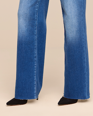 Person wearing blue wide-leg jeans and black pointed-toe heels. The image is cropped to show only the legs and feet, with a plain beige background.