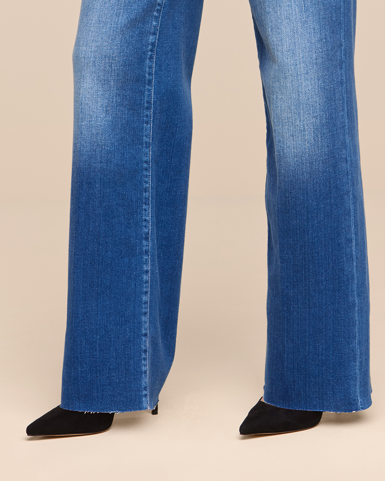 Person wearing blue wide-leg jeans and black pointed-toe heels. The image is cropped to show only the legs and feet, with a plain beige background.