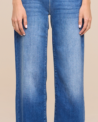 A person wearing blue, straight-leg jeans stands with their hands at their sides against a beige background. Only the lower torso and upper legs are visible.