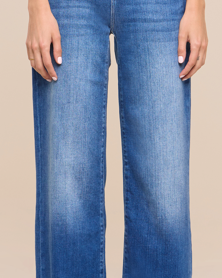 A person wearing blue, straight-leg jeans stands with their hands at their sides against a beige background. Only the lower torso and upper legs are visible.
