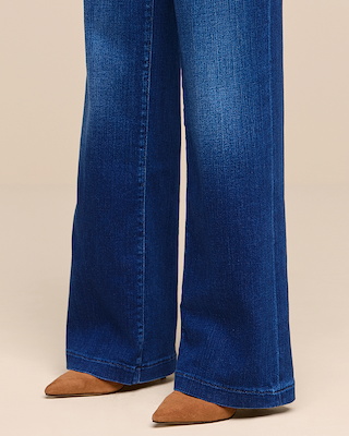 Person wearing wide-leg blue jeans and brown pointed-toe shoes, standing on a neutral tan floor with a plain beige background.