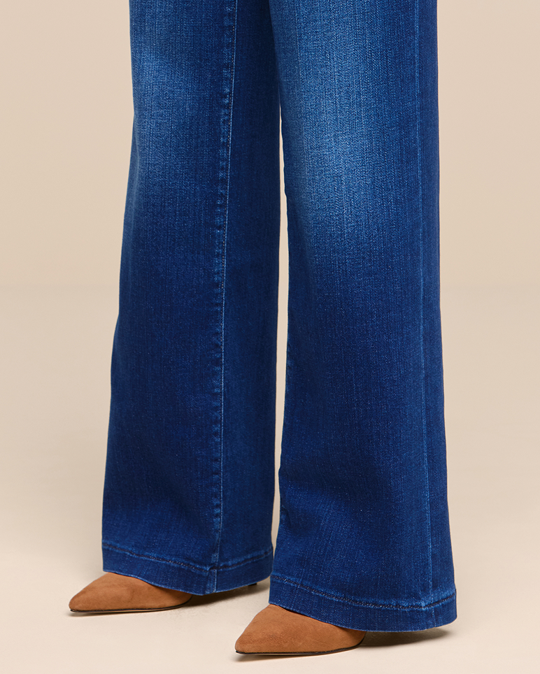 Person wearing wide-leg blue jeans and brown pointed-toe shoes, standing on a neutral tan floor with a plain beige background.