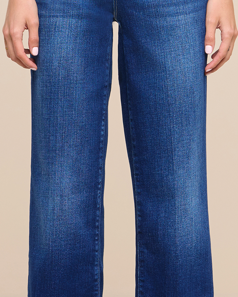 A person wearing blue denim jeans stands against a plain beige background, with their hands relaxed at their sides and fingernails painted white.