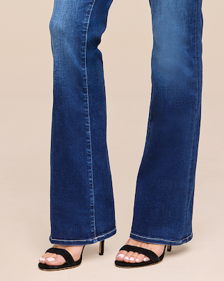 A person wearing blue bootcut jeans and black open-toe heels stands on a beige floor. Only the legs and feet are visible.