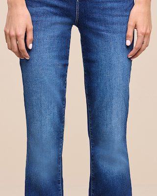 Person standing with arms relaxed at sides, wearing blue jeans and light pink nail polish. The image is cropped to show the lower torso, legs, and hands against a plain beige background.