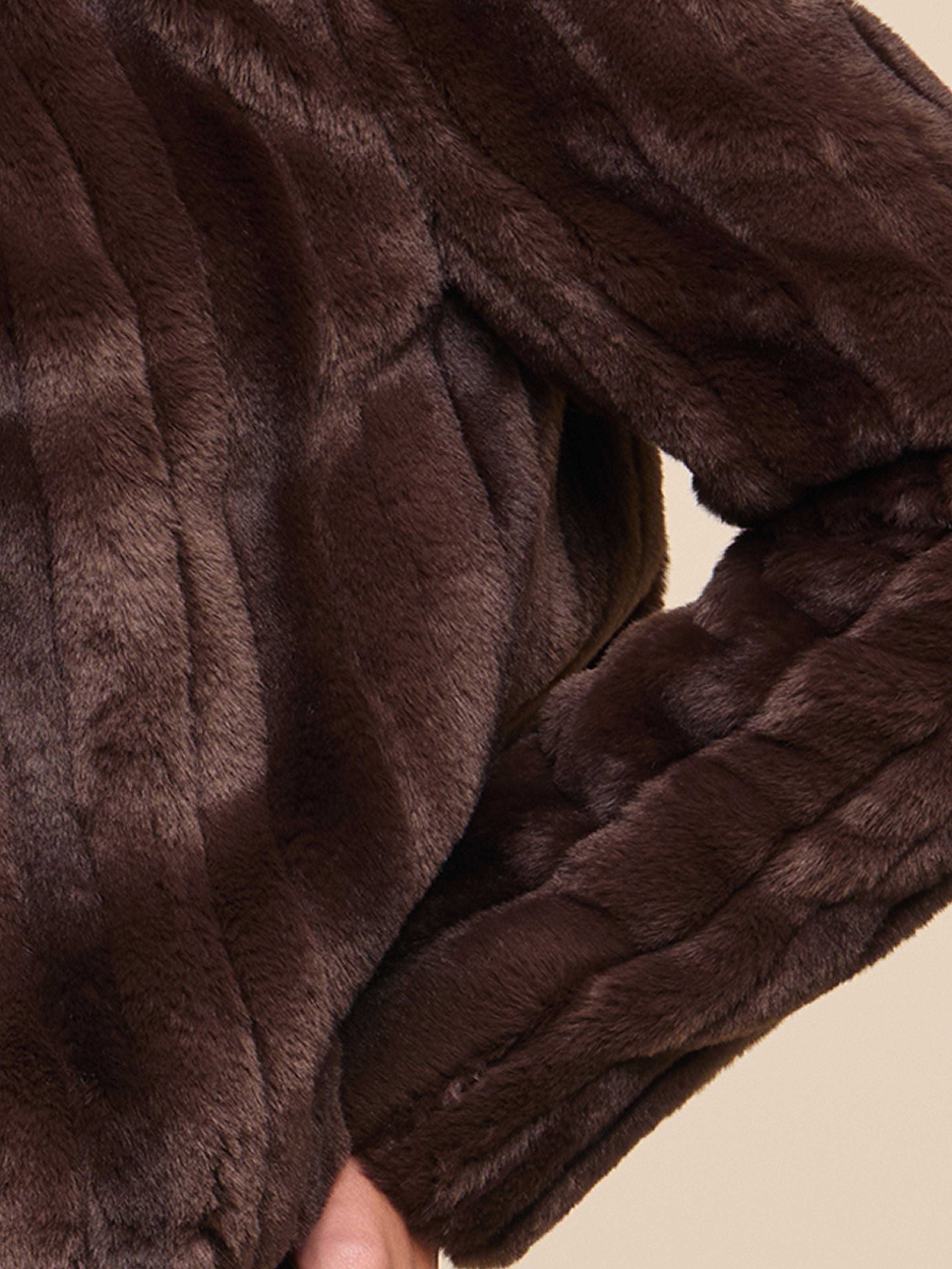 A close-up of a person wearing a brown faux fur coat, with one arm bent and hand resting on their hip.