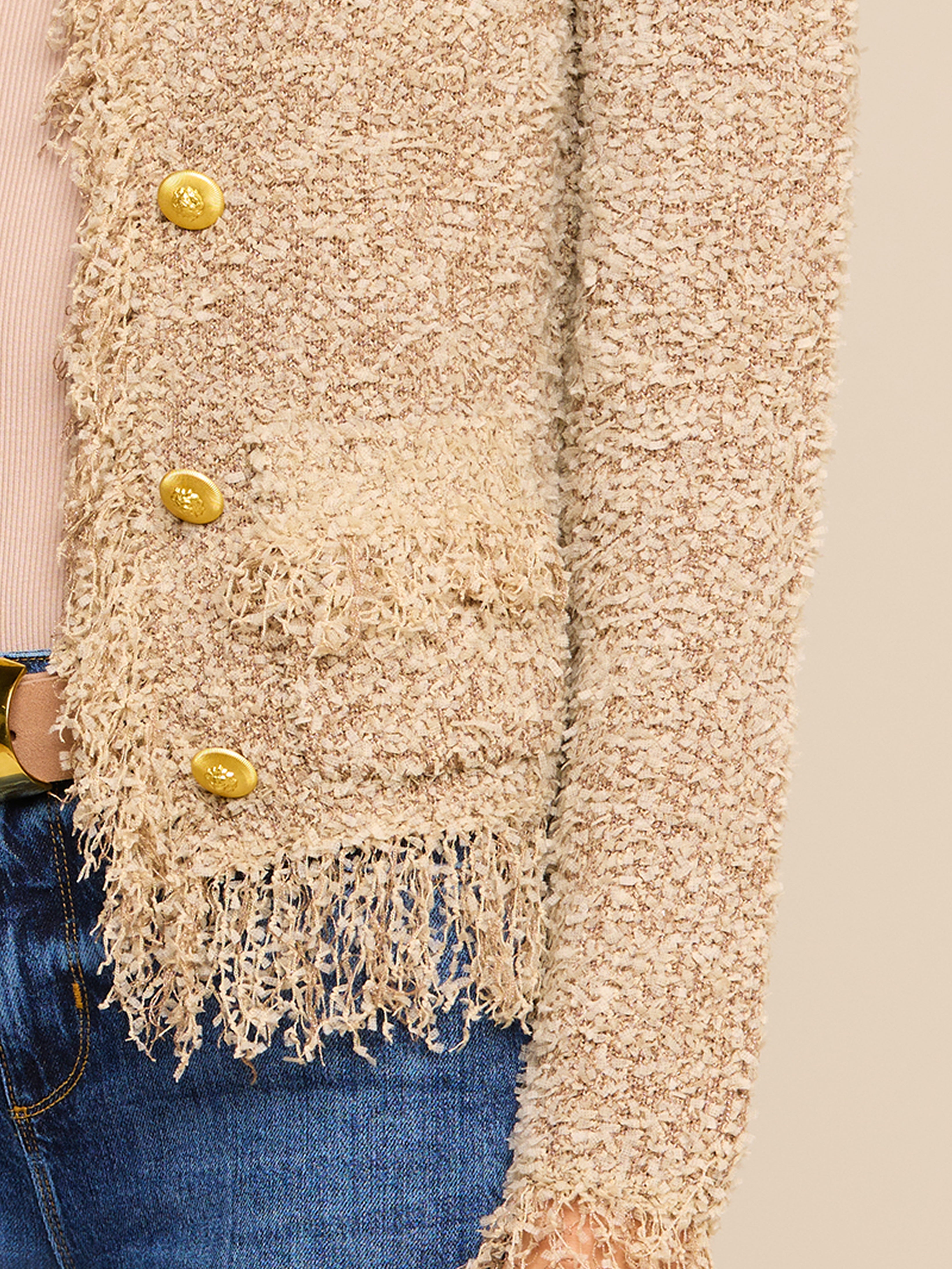 A close-up of a textured beige jacket with gold buttons and fringe details at the hem, paired with blue jeans and a light pink top.