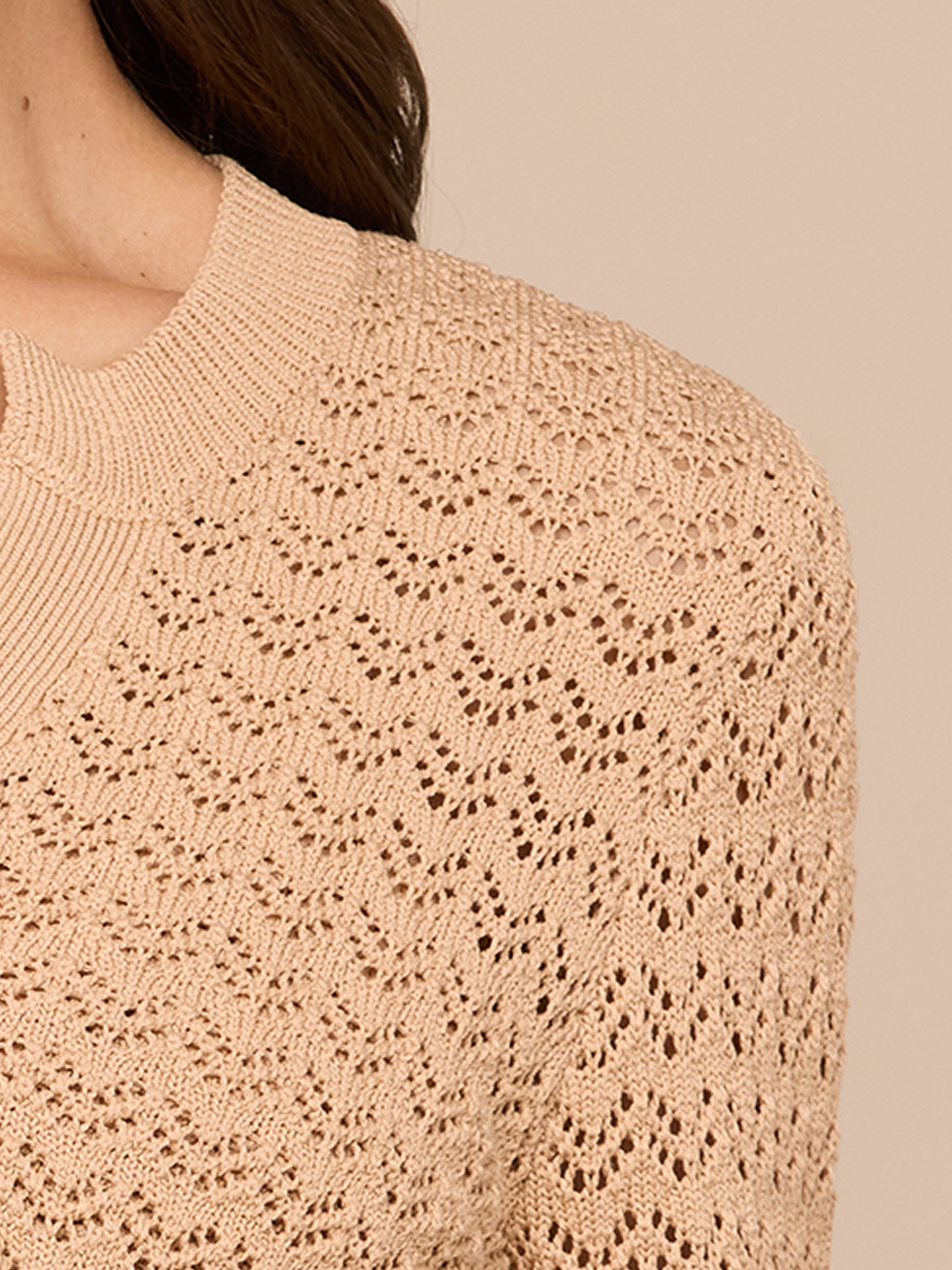 Close-up of a person wearing a beige, knitted sweater with a textured, lace-like pattern. Only part of the shoulder, upper chest, and neck is visible; background is a similar beige color.
