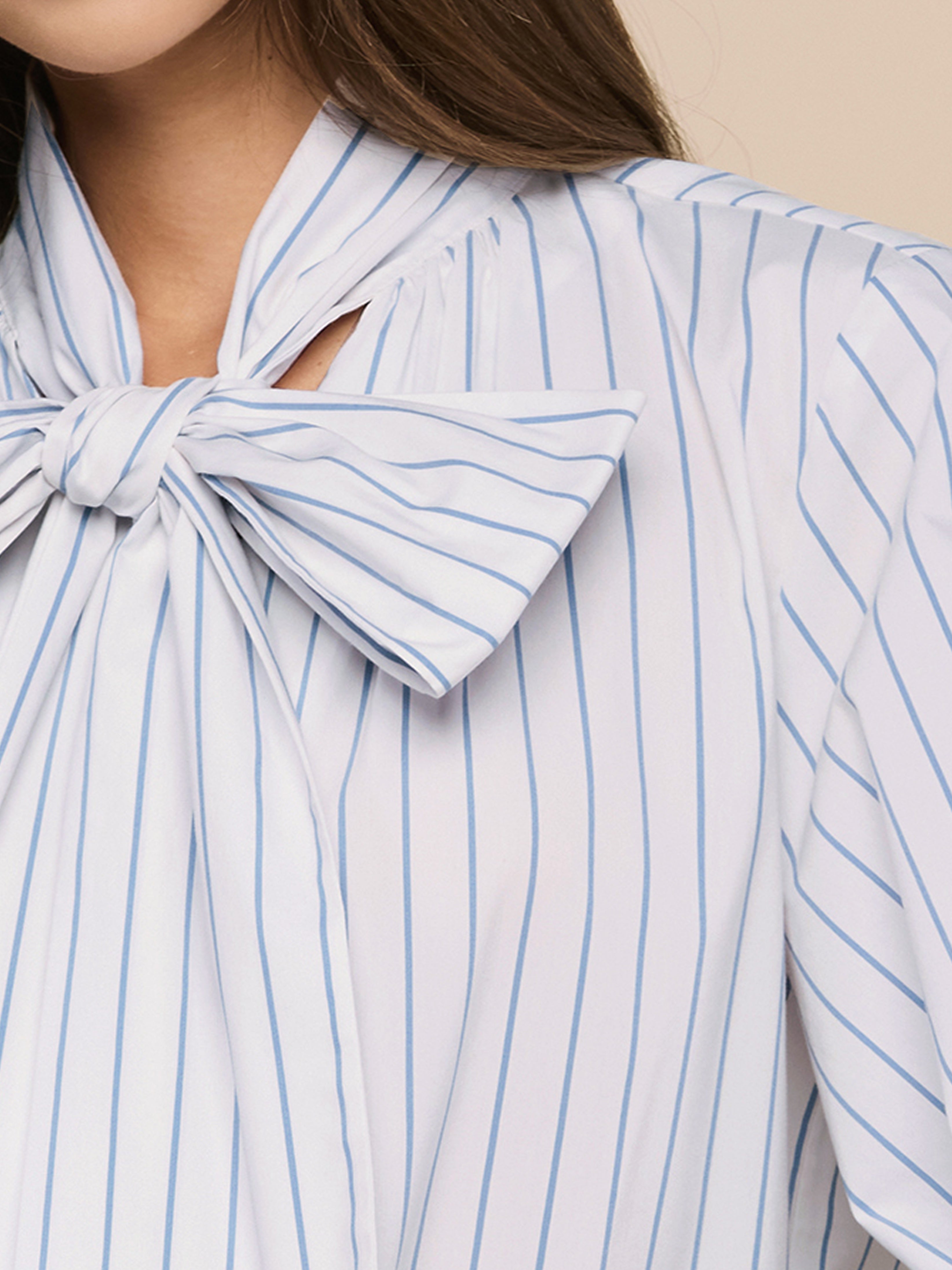 Close-up of a woman wearing a white blouse with light blue vertical stripes and a large bow tied at the collar. Only her lower face, neck, and part of her hair and shoulder are visible.