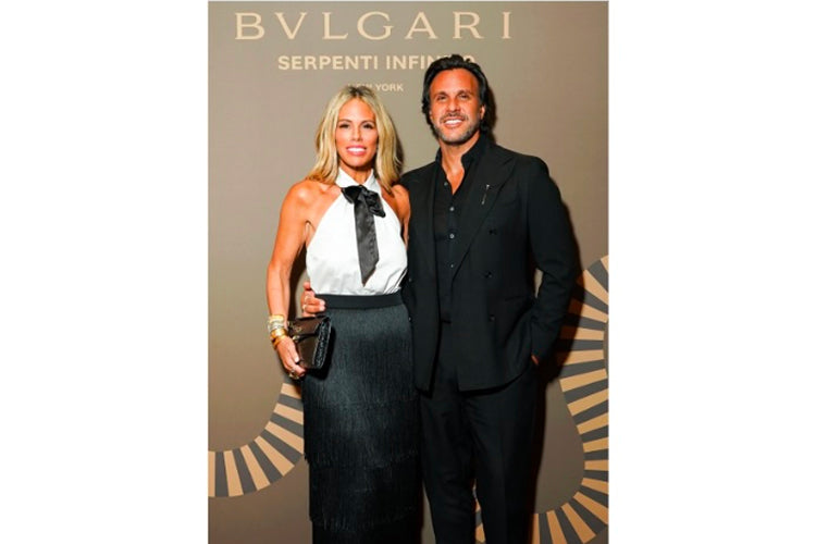 A woman in a white halter top and black skirt poses next to a man in a black suit in front of a Bvlgari Serpenti backdrop.