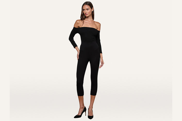 A woman in a black off-the-shoulder midi jumpsuit paired with black pointed-toe heels.