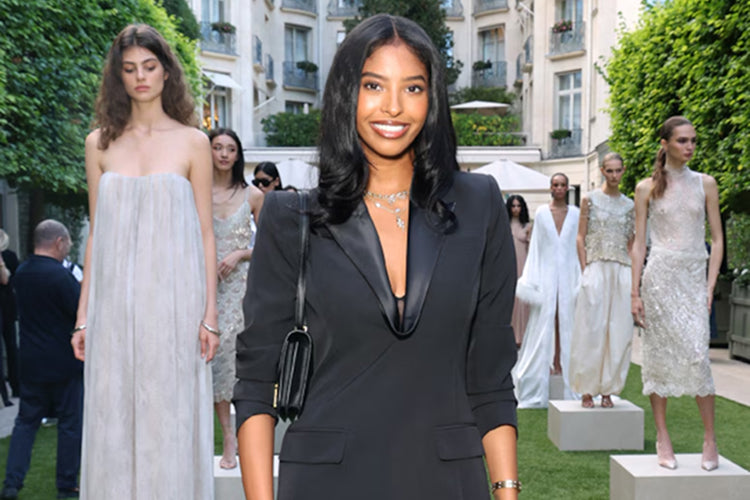 Natalia Bryant smiles while wearing a black blazer in the foreground, with models in white and cream outfits standing on pedestals in the background.