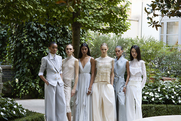 Six models stand in a line outdoors in a garden, wearing various styles of luxury neutral-toned clothing.