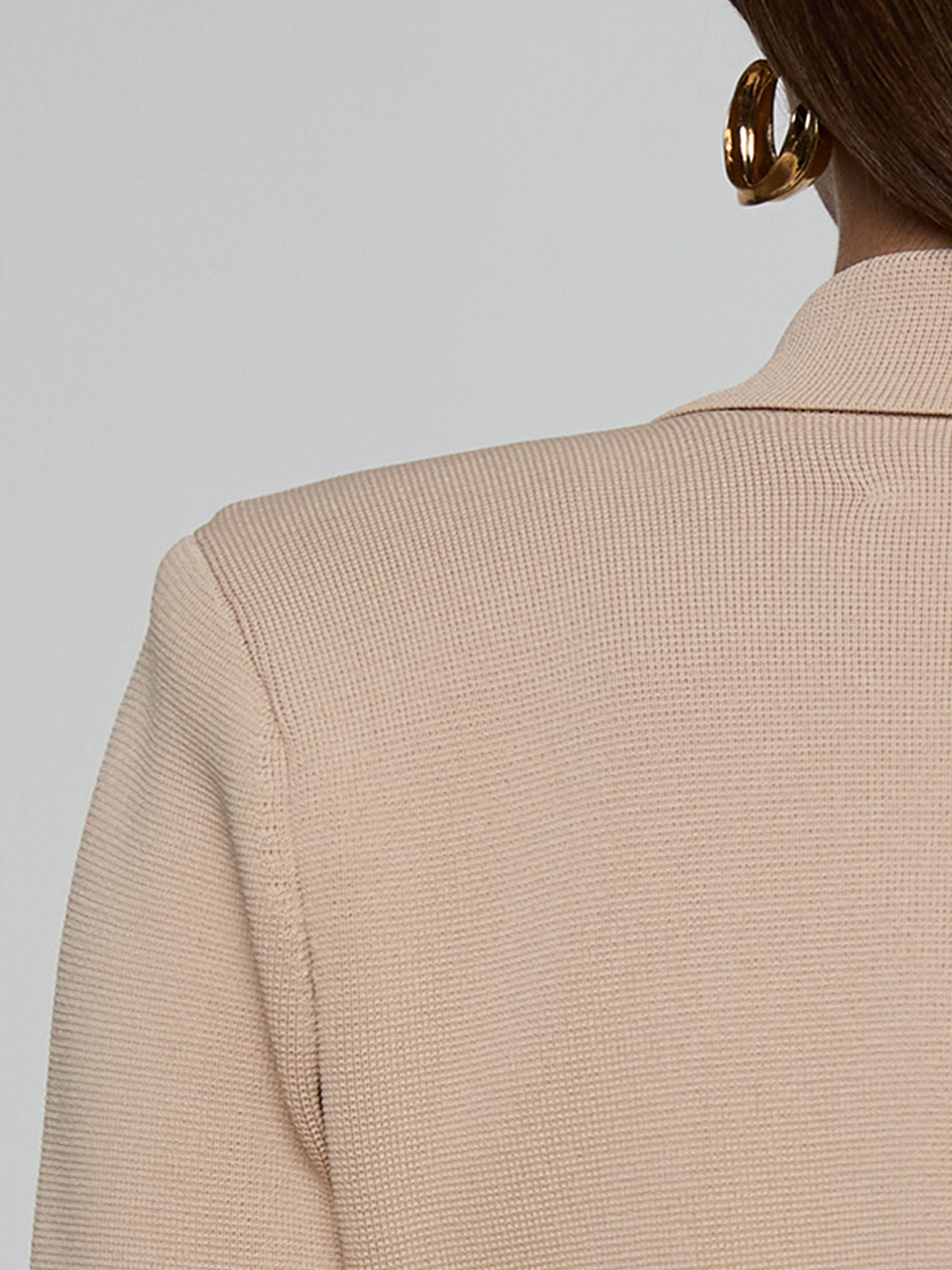 Close-up of a person wearing a textured beige blazer and large gold hoop earrings, with only the back and shoulder visible against a plain light background.