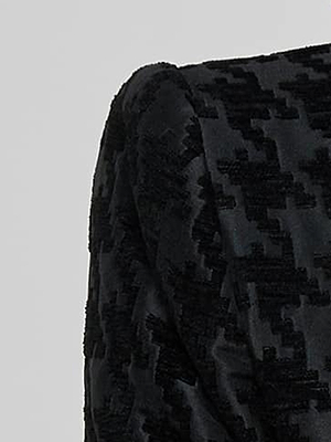 Close-up of a black textured fabric with a raised geometric pattern, possibly part of a garment such as a jacket or dress sleeve, set against a plain light gray background.