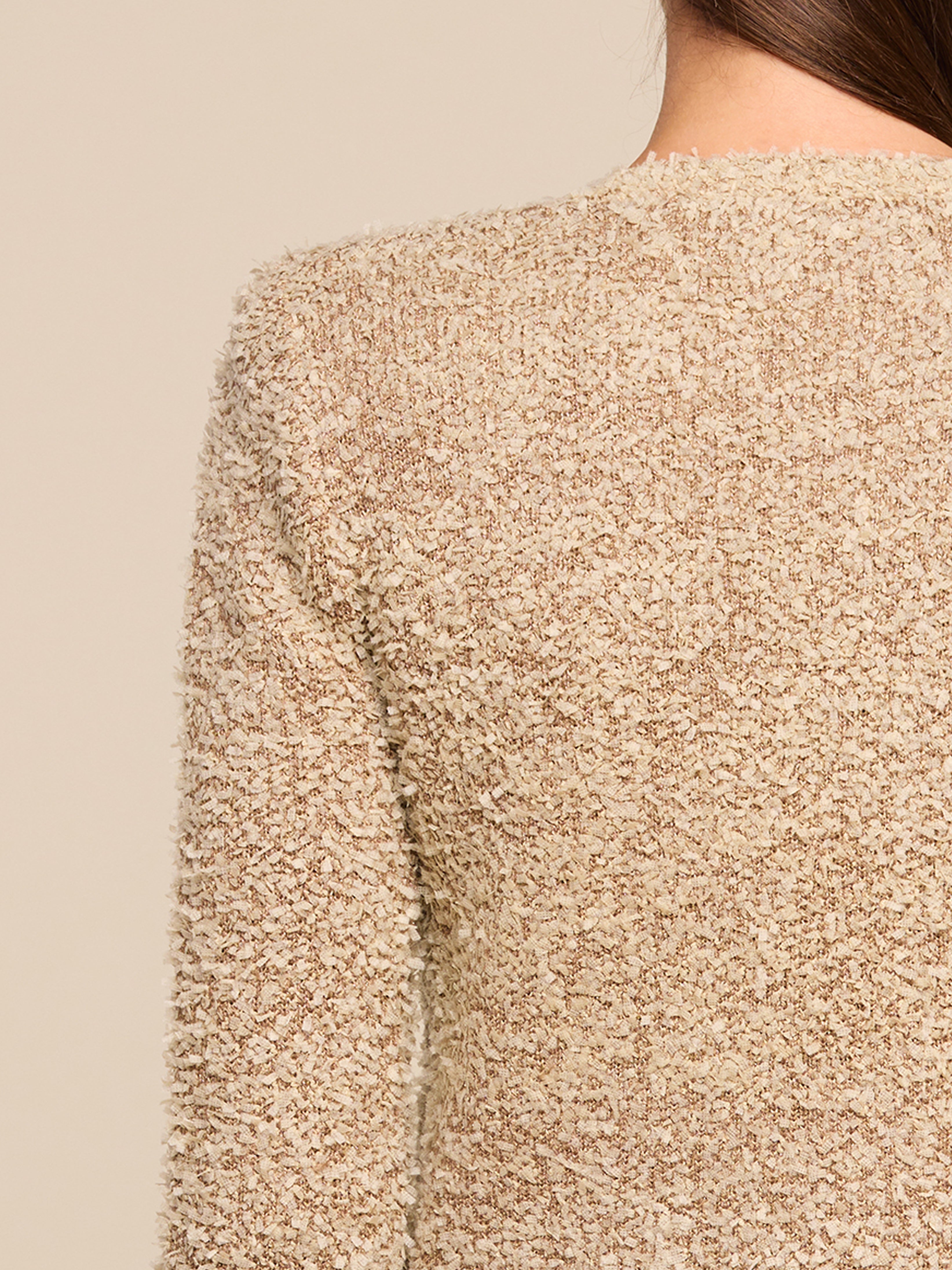 Close-up of a persons upper back and shoulder, wearing a textured, beige, fuzzy sweater. Only part of the head with long brown hair and neck is visible; background is a plain, neutral color.