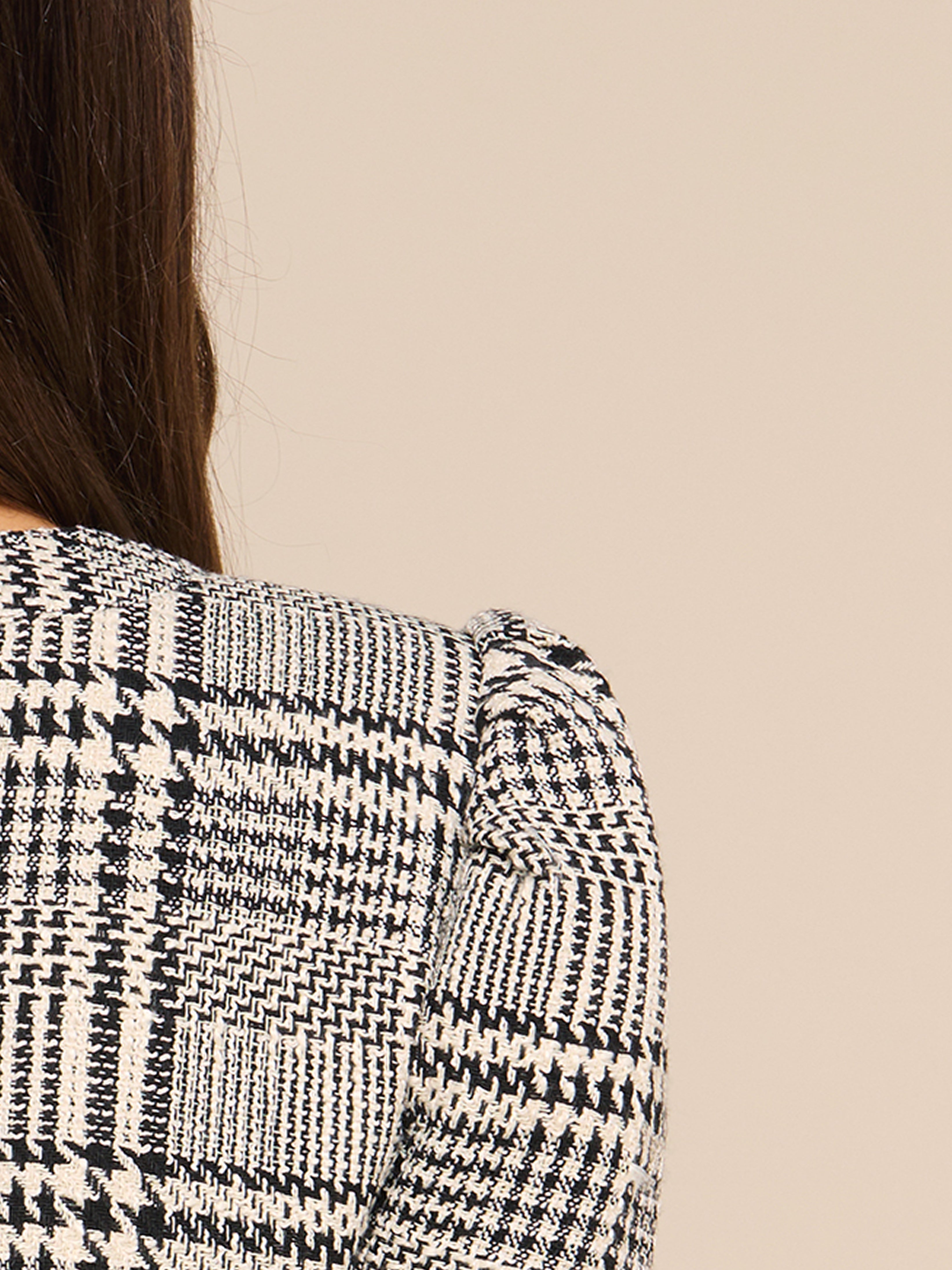 The image shows the back view of a person wearing a black-and-white plaid textured jacket. Only the shoulder and partial back are visible, with long brown hair draped over one side against a plain beige background.