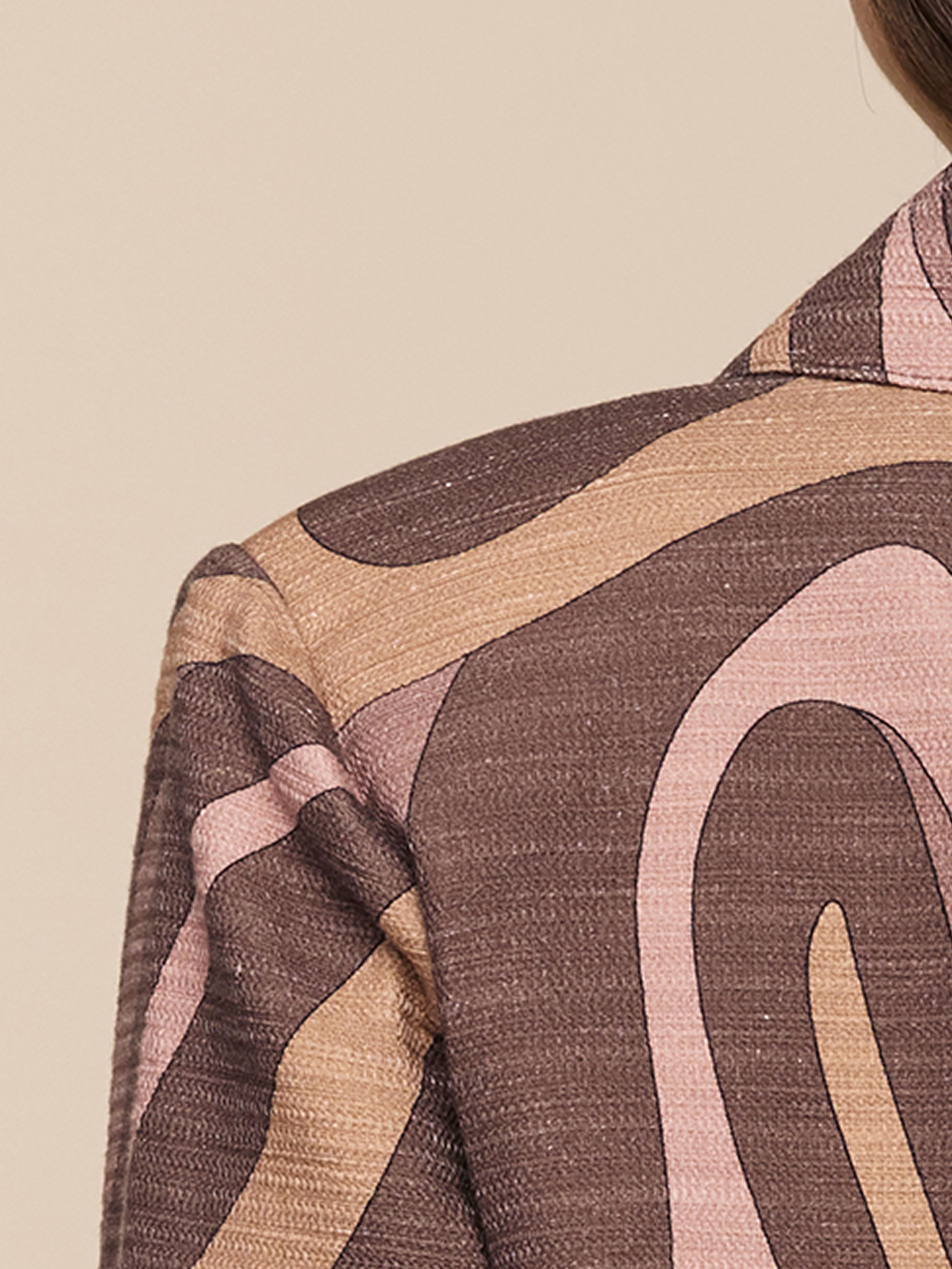 Close-up of a persons shoulder wearing a brown jacket with beige and light pink abstract, wavy patterns on it; neutral beige background.