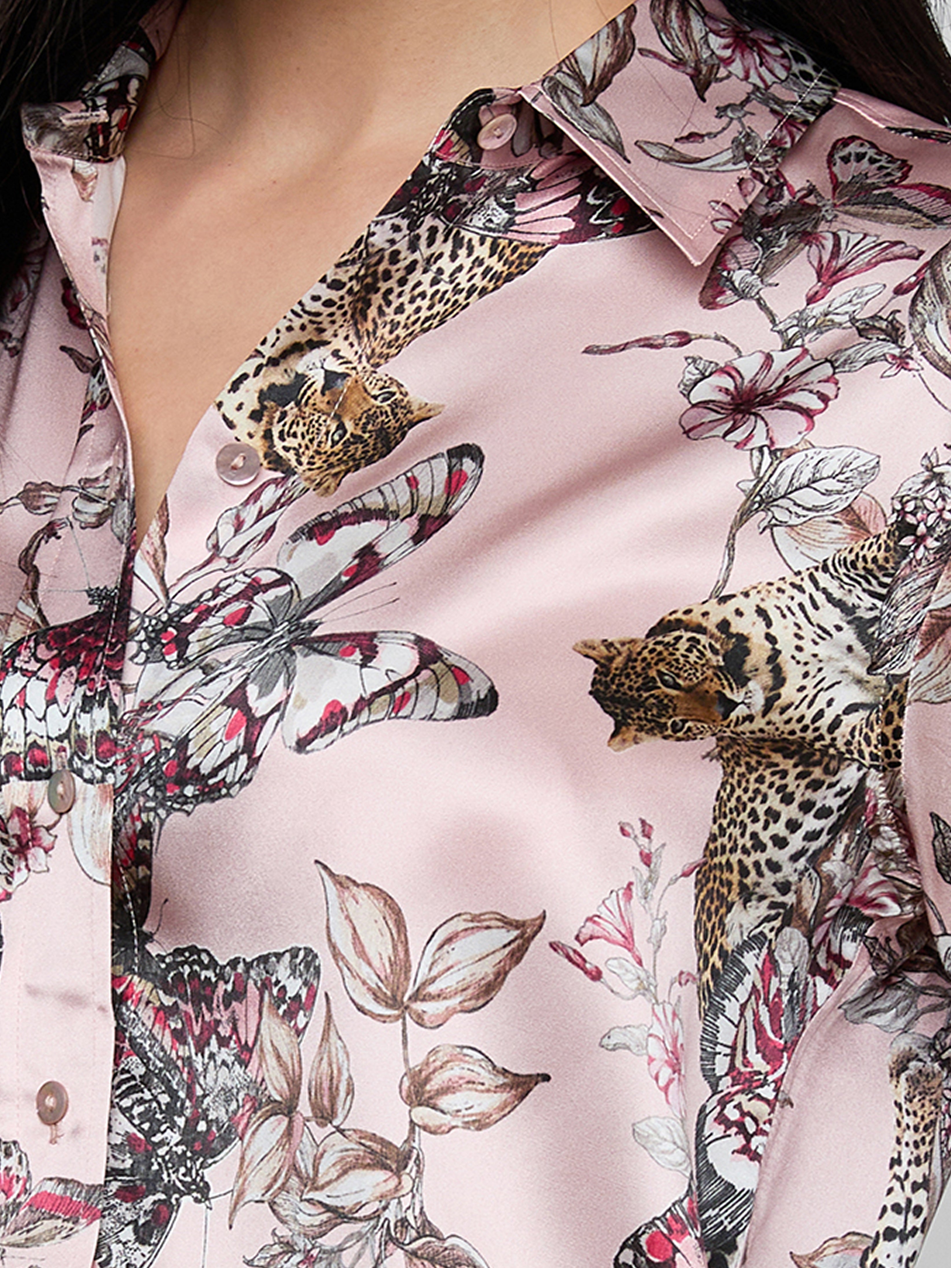 A close-up of a person wearing a silky, light pink shirt with a pattern featuring leopards, butterflies, and flowers in shades of black, white, and pink. Only the lower face, neck, and part of the shirt are visible.