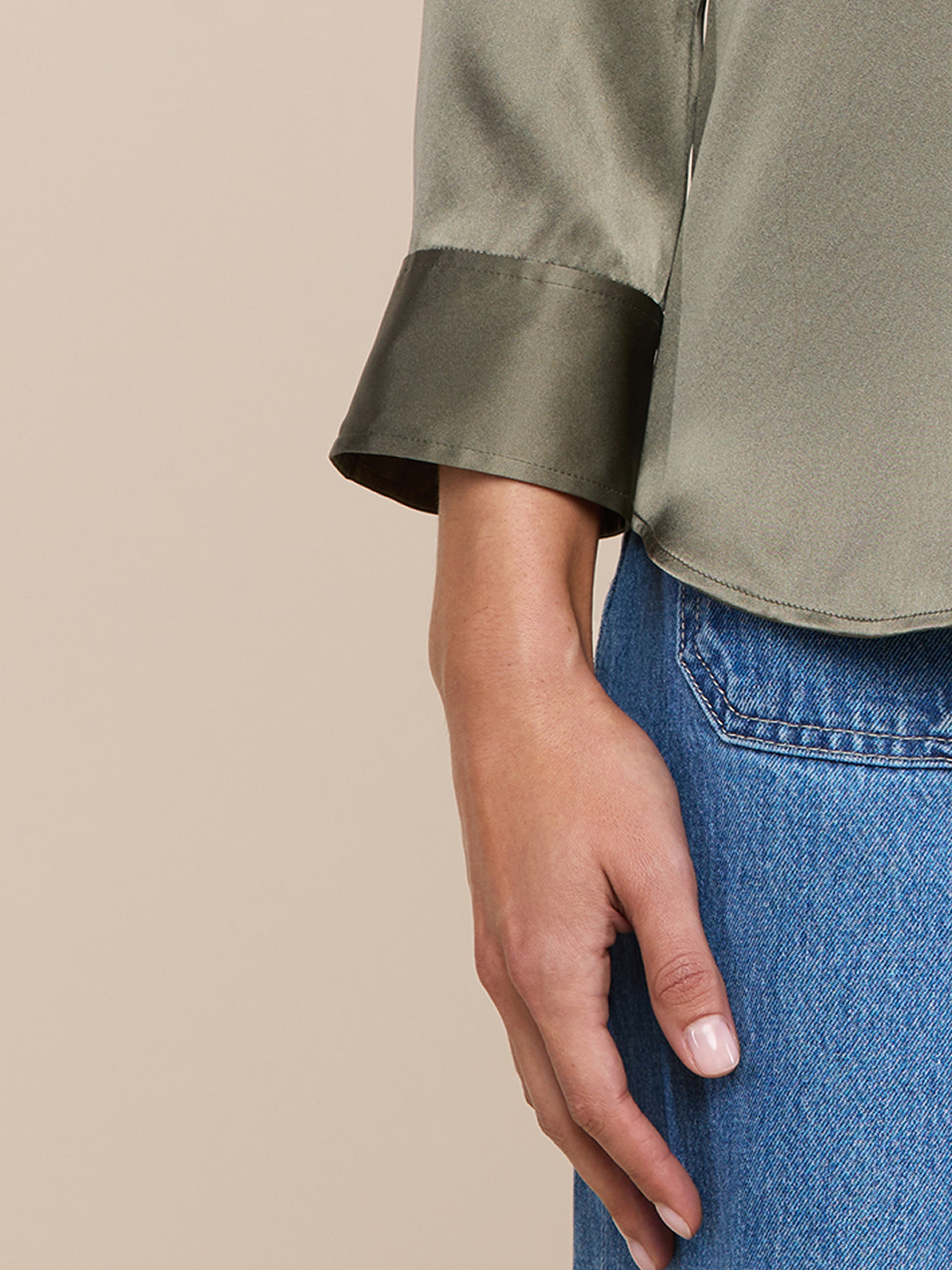 A close-up of a persons left hand resting by their side, wearing a shiny olive-green long-sleeve blouse with wide cuffs and blue jeans against a plain beige background.