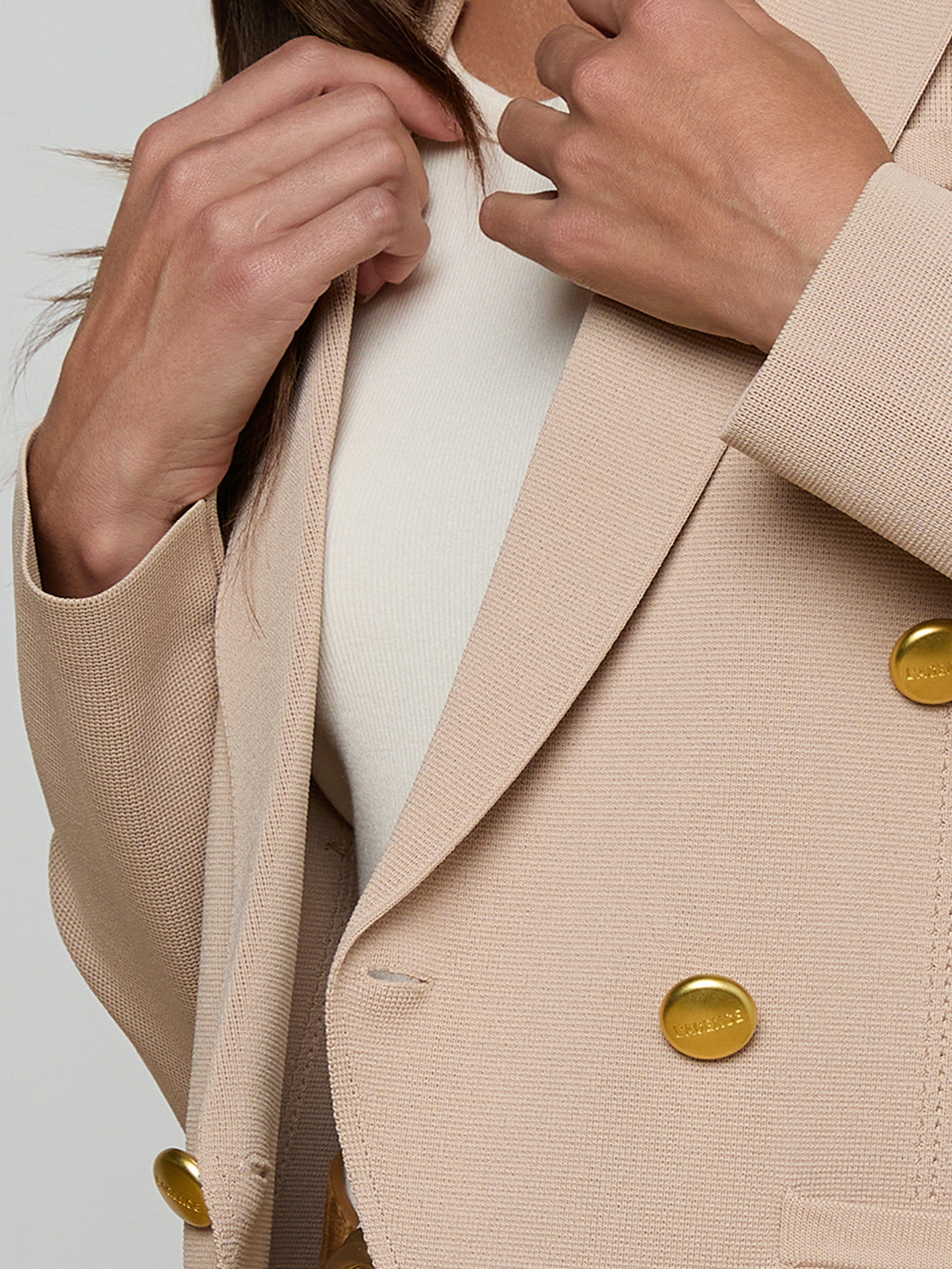 A person wearing a cream top and a beige blazer with large gold buttons is adjusting the lapels of the blazer with both hands.
