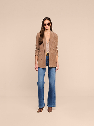 A woman stands against a beige background wearing sunglasses, the tan L’AGENCE Arabell Romper, a belt, animal print heels, and blue jeans. Her long hair is styled in loose waves.