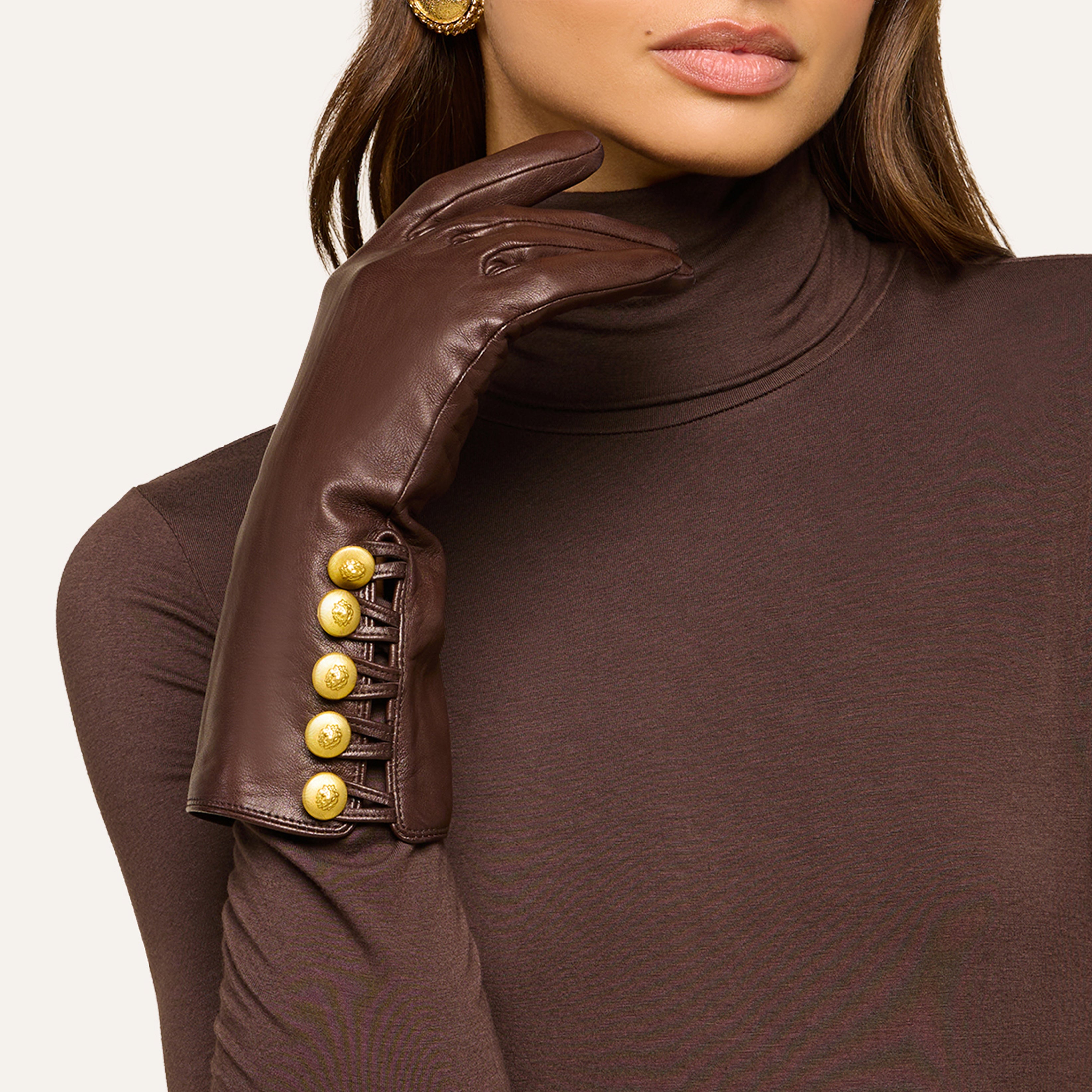 A woman in a brown turtleneck wears dark brown leather gloves with gold buttons, holding one gloved hand near her chin. Only her lower face is visible, highlighting the elegance of the glossy leather against her skin.