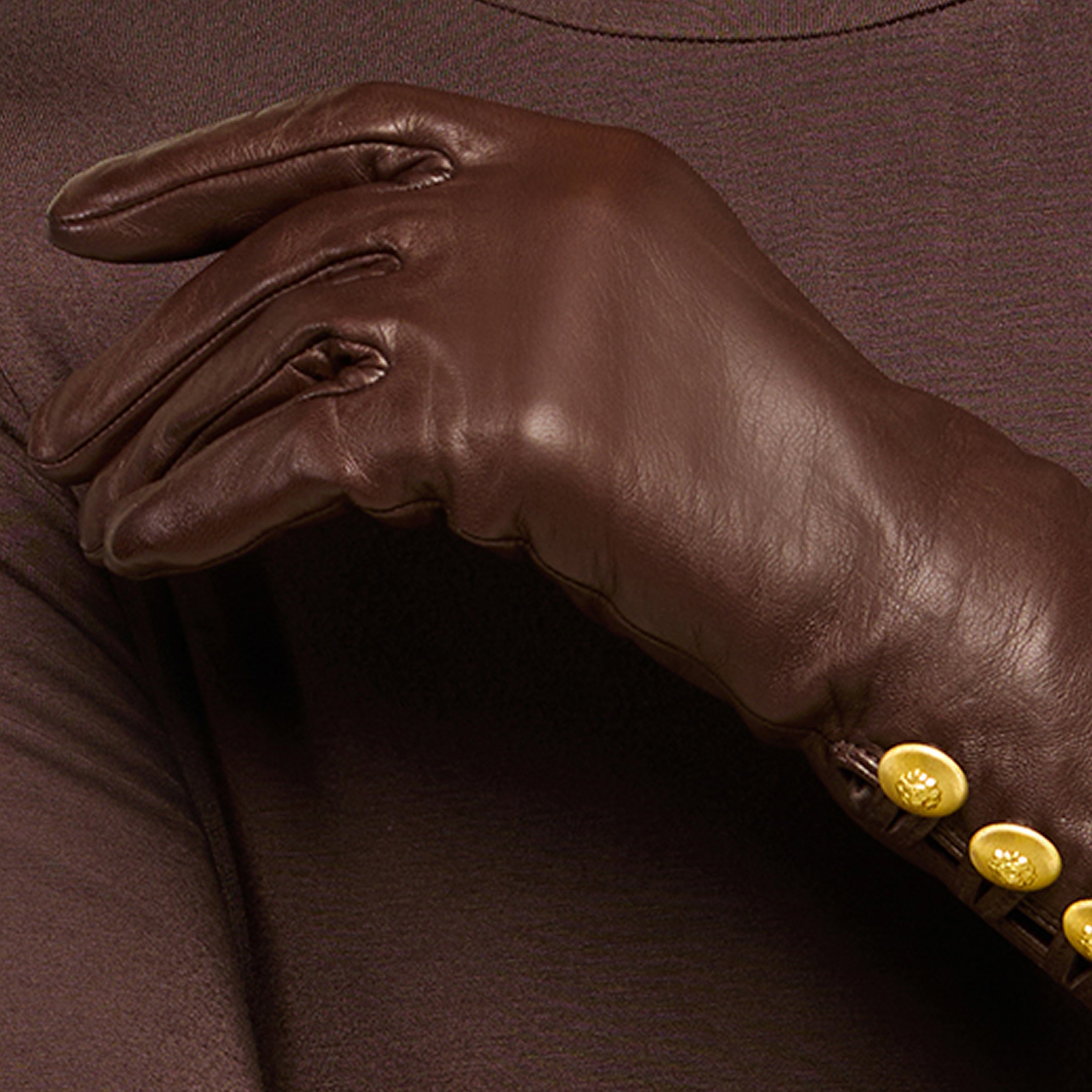 A hand wearing a brown leather glove with three yellow buttons rests on a brown tweed fabric background.