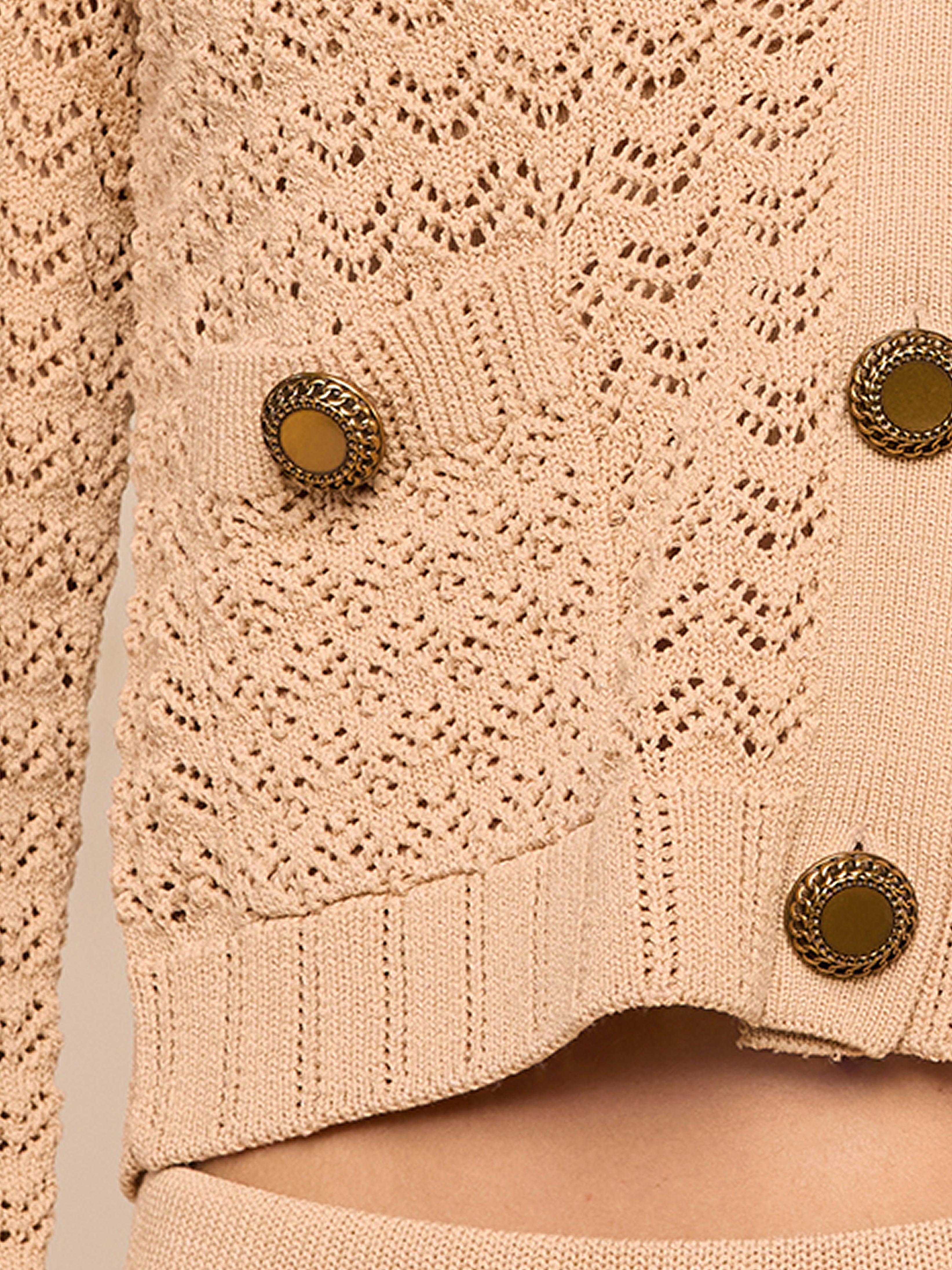 Close-up of a beige knit cardigan with a lacy pattern, featuring round gold buttons and a front pocket. The edge of the cardigan rests above matching knit bottoms.