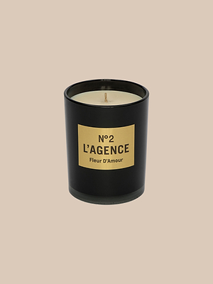 A black candle jar with beige soy-blend wax and a centered wick, labeled “Fleur DAmour” in black on a gold rectangular label, is set against a plain beige background.