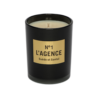 A black candle jar with a gold rectangular label reading “Suède et Santal,” featuring a single wick and hand-poured soy wax, is photographed on a white background.