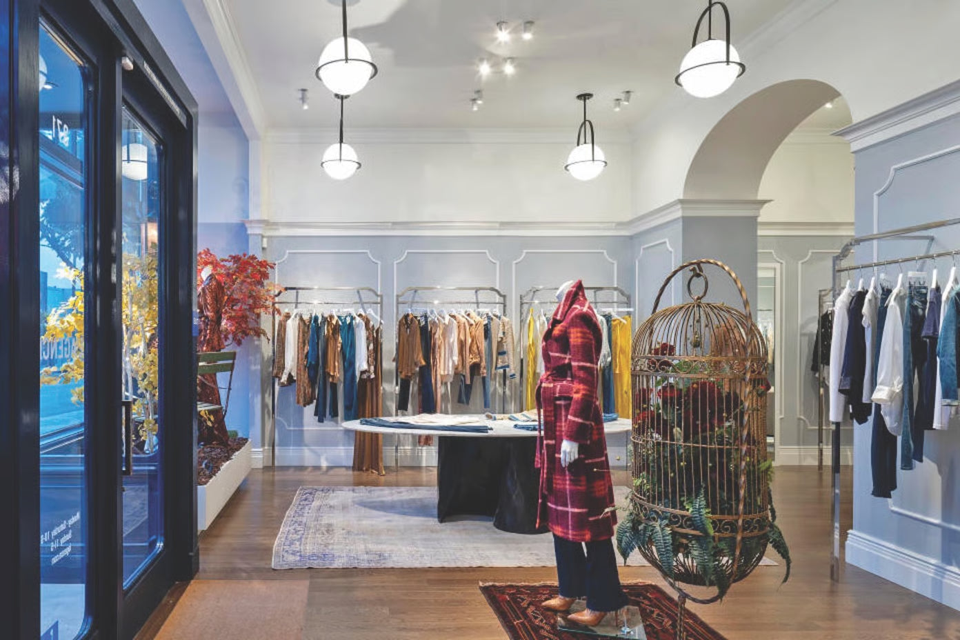Chic clothing boutique interior with racks of clothes, a table, decorative lighting, and a mannequin in a red plaid coat. A vintage birdcage with greenery sits near the entrance.