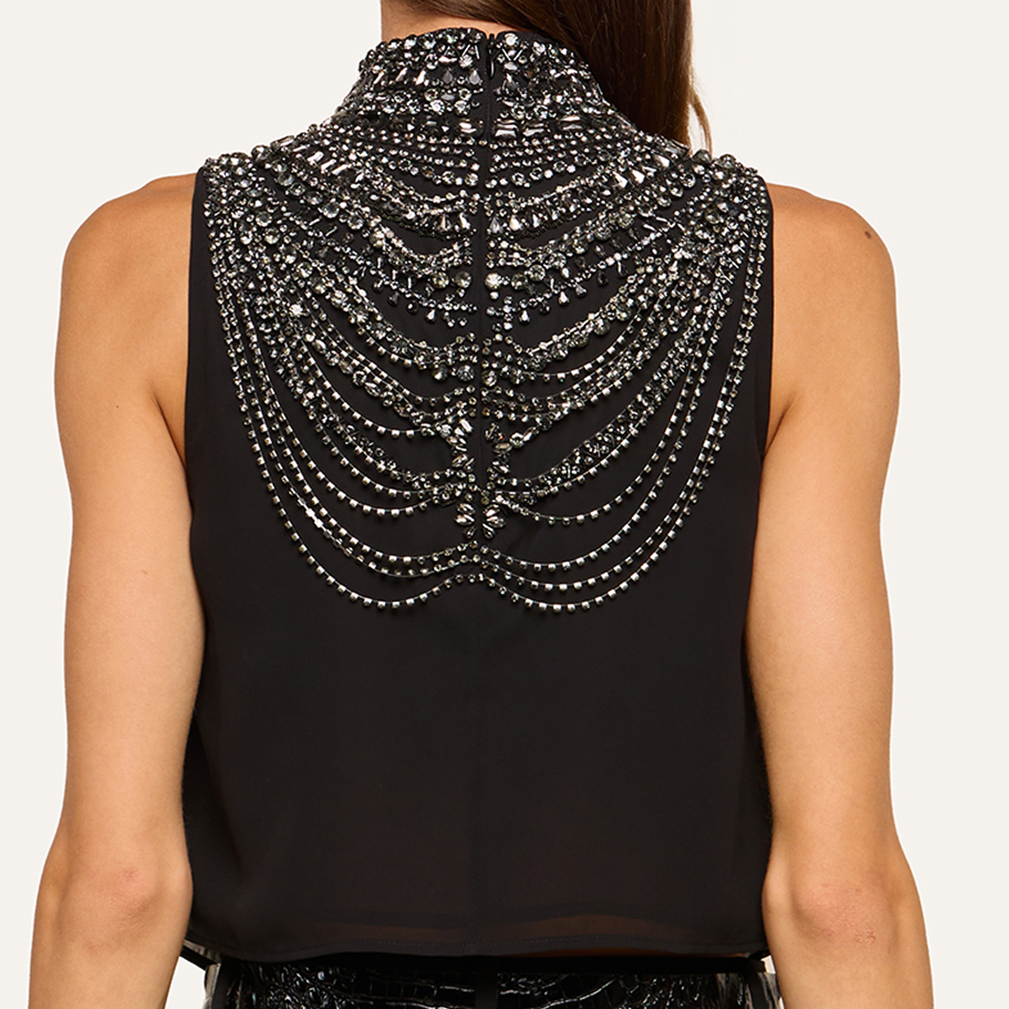 A woman is dressed in a sleeveless black top, featuring intricate, shiny silver bead embellishments that form elegant curved patterns across the back.