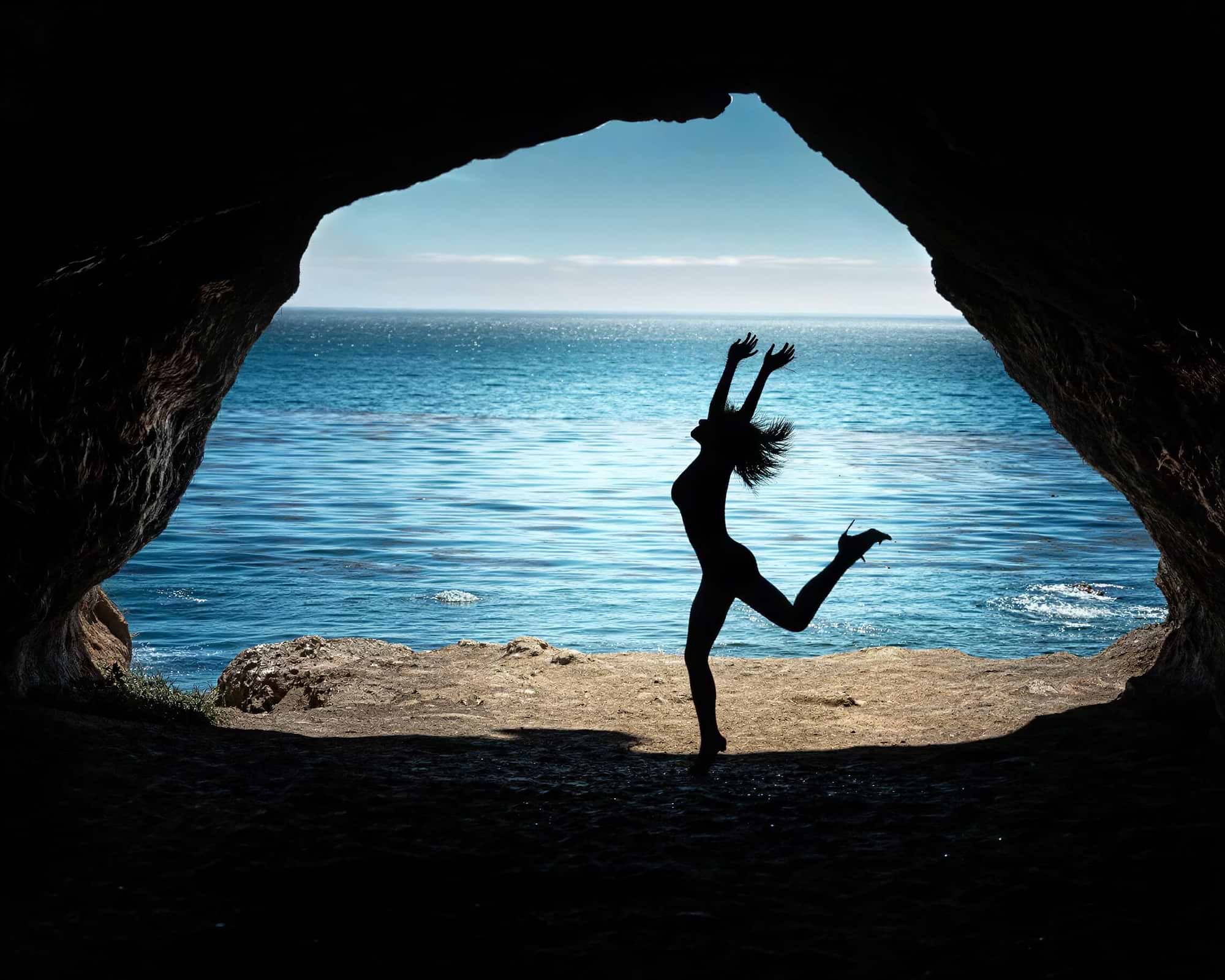 A silhouetted person jumping in front of a cave opening that frames a bright blue ocean scene, creating a dramatic high-contrast silhouette effect.