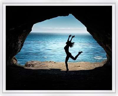 A framed photographic print showing a silhouetted dancer jumping in front of a cave opening that reveals a bright blue ocean and sky.